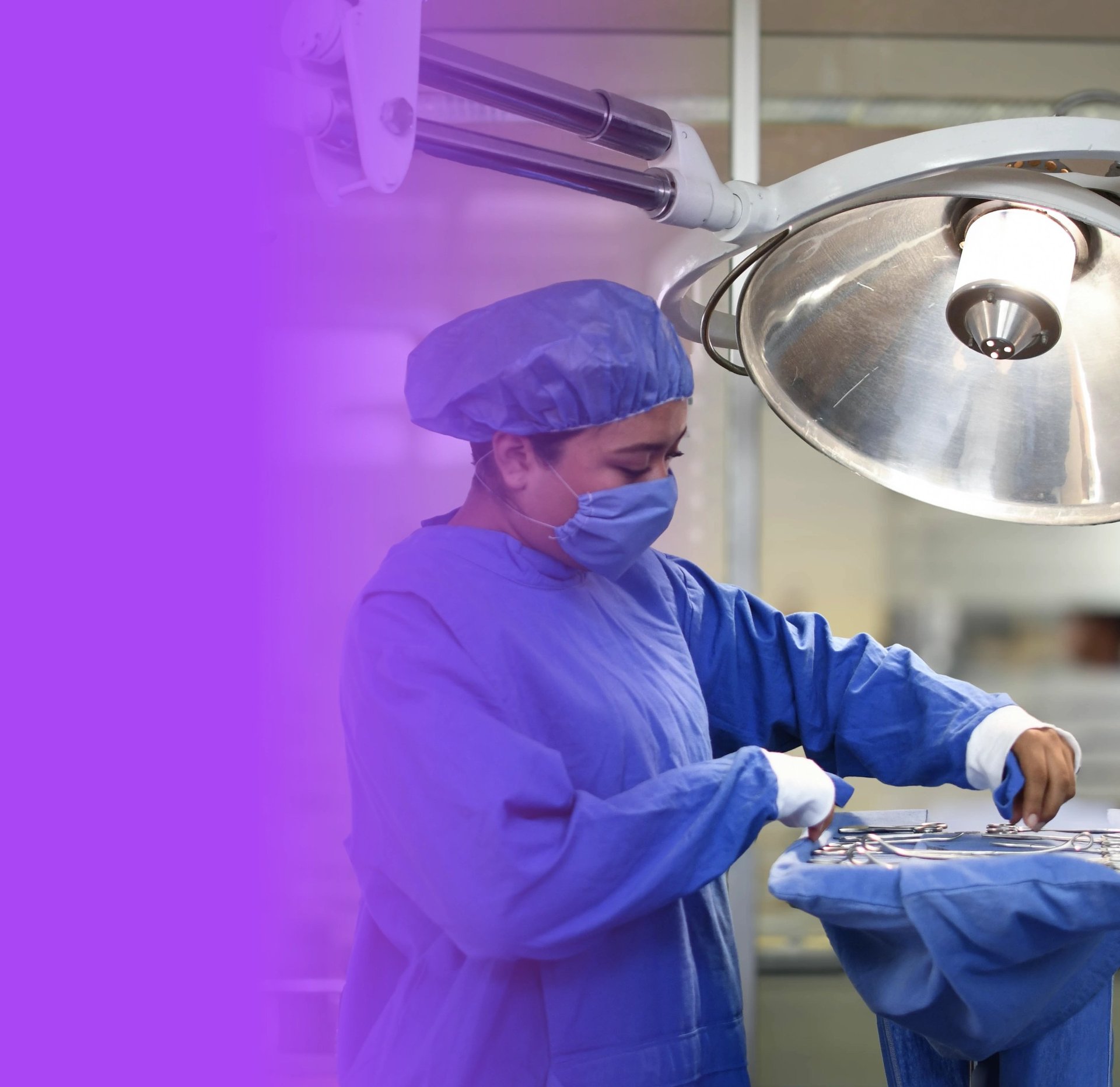 Surgeon in blue scrubs and surgical cap preparing sterile instruments under an overhead surgical light