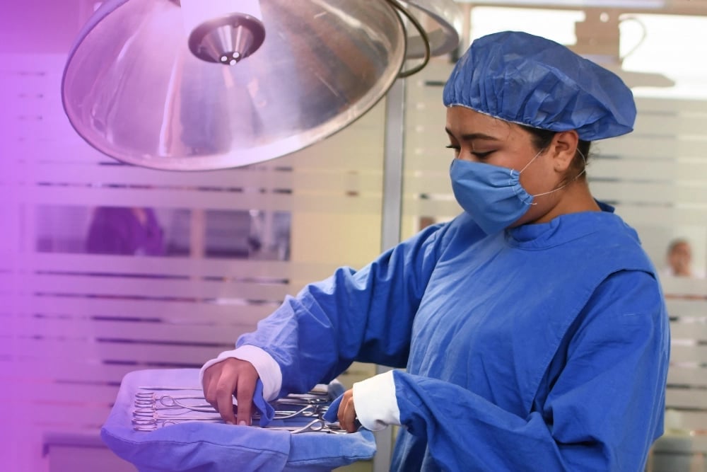 Surgeon in blue scrubs and mask prepares surgical instruments under purple lighting in operating room
