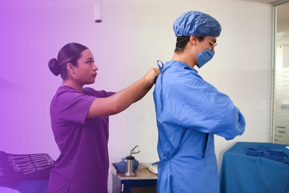 Healthcare worker in purple scrubs helping patient in blue surgical gown and cap put on protective equipment in medical facility