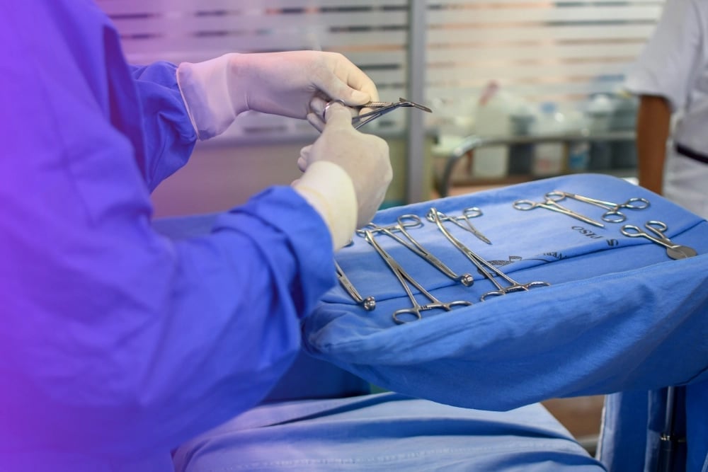Surgeon in purple scrubs holding surgical instrument over tray of sterilized surgical tools in operating room