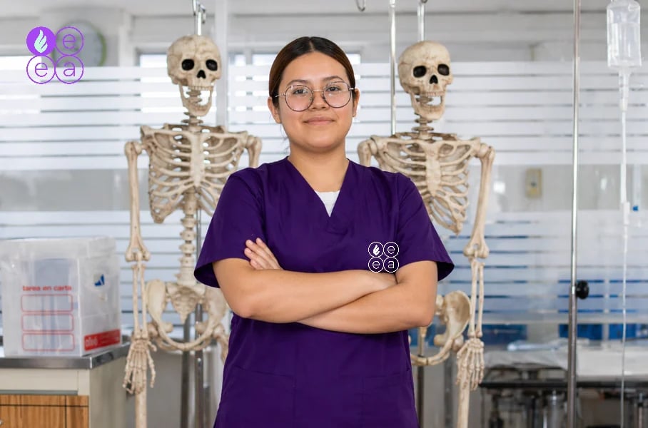 Female healthcare worker in purple scrubs standing with arms crossed in front of two skeleton models in a medical office