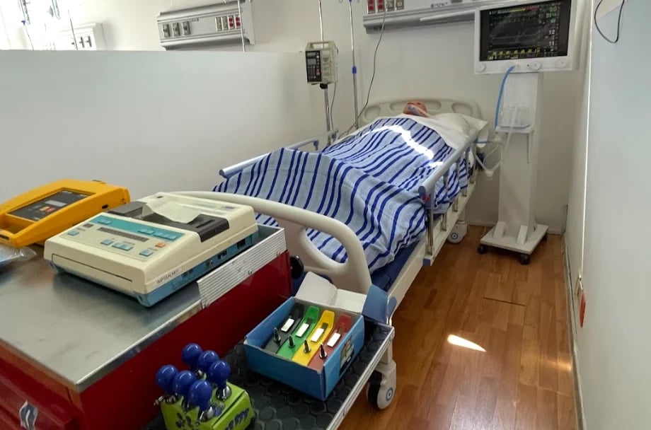 Hospital room with medical training mannequin on bed, monitoring equipment on walls, and medical supplies on table