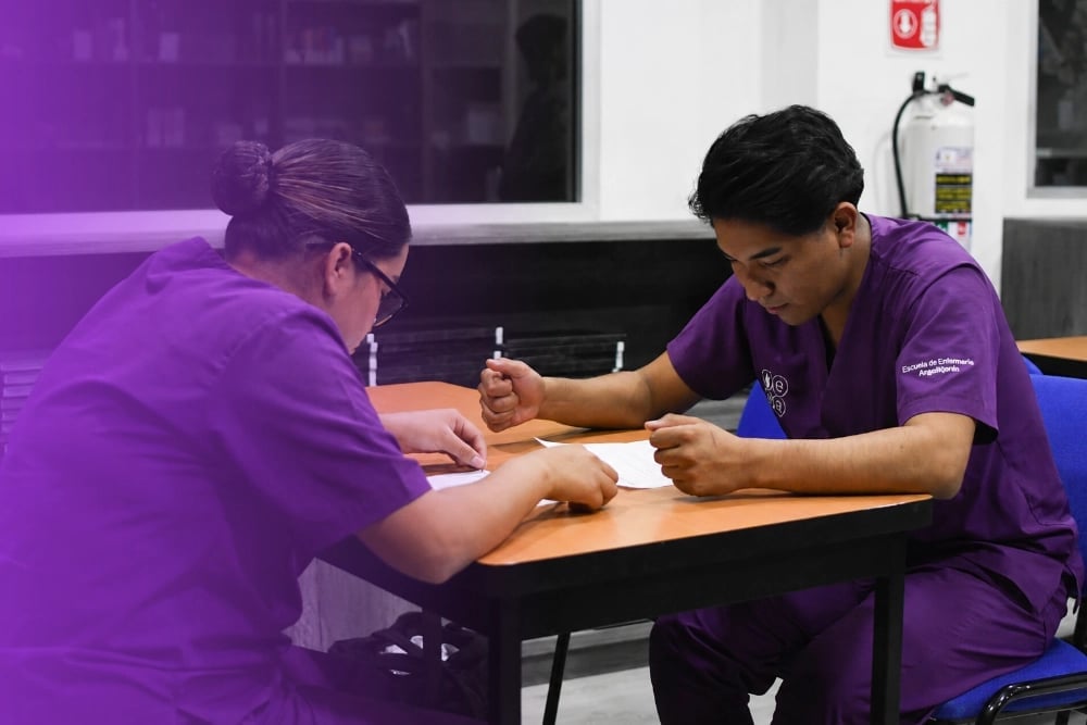 Two people in purple shirts sitting at a table, one writing while the other observes during what appears to be a training or instructional session