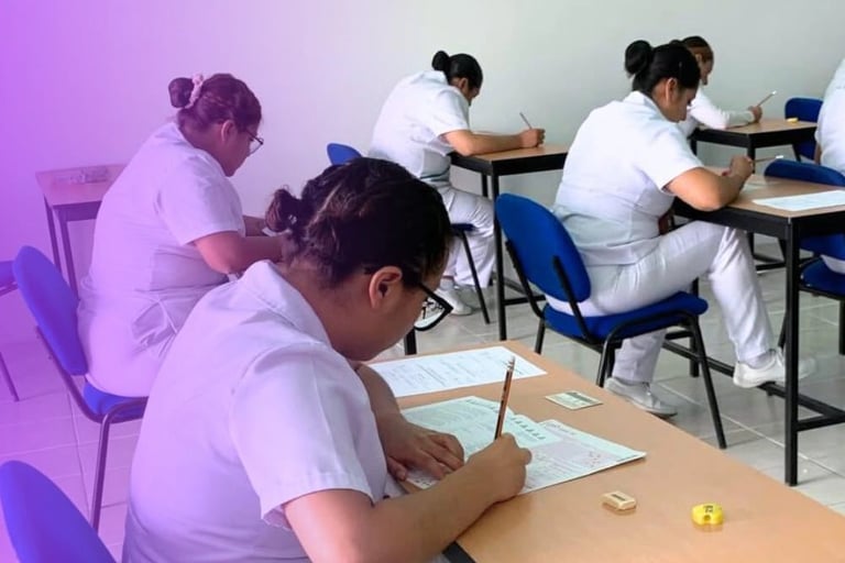 Students in white uniforms taking an exam at individual desks in a classroom with blue chairs